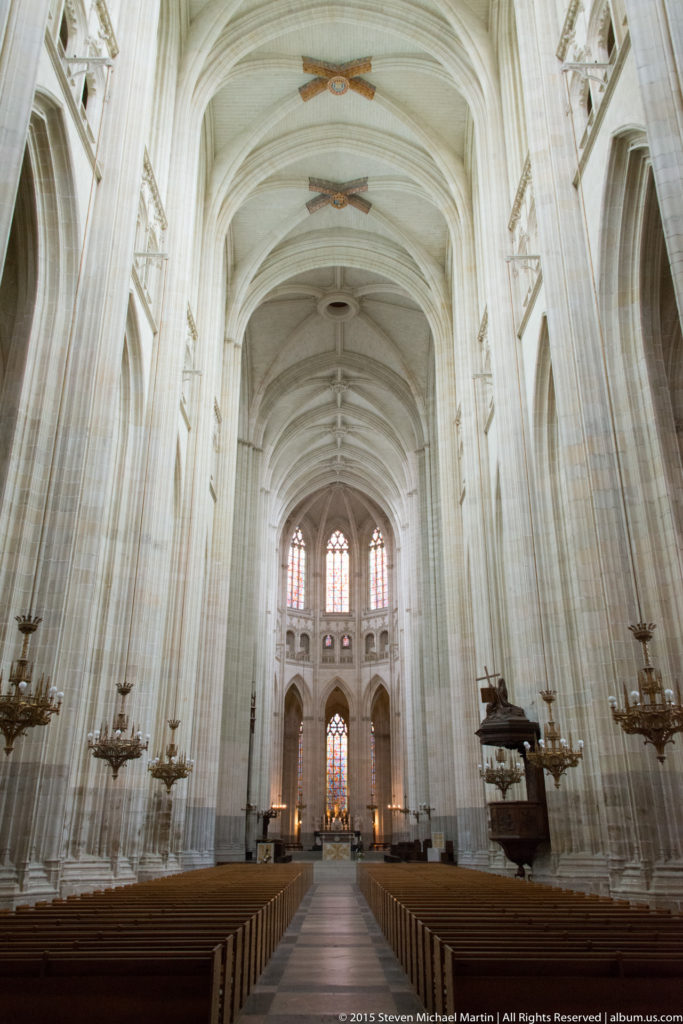 Symmetrical view down Nantes Cathedral's nave with rows of pews, pale Gothic piers, ribbed vaults, and distant stained glass.