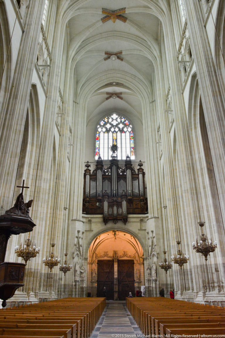 Great pipe organ on a high gallery at the west end of Nantes Cathedral, framed by pale stone piers and stained glass.