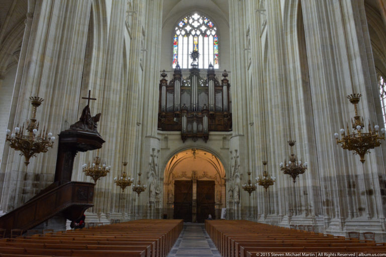 Nantes Cathedral interior with the great organ above the west entrance, carved pulpit at left, chandeliers, pews, and tall piers.