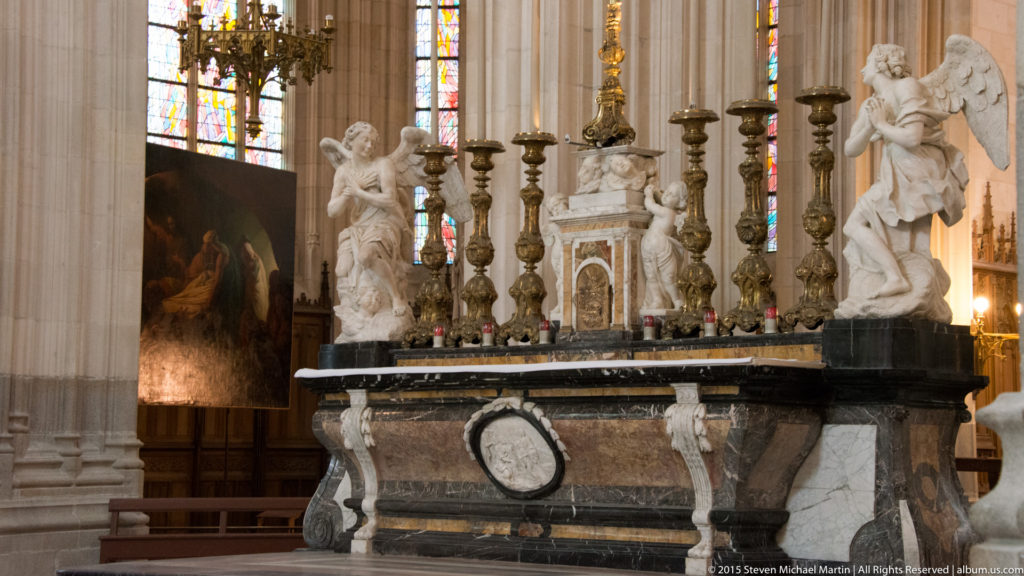 Side view of a dark marble cathedral altar with white angels, tall candlesticks, an oval medallion, painting, and stained glass.