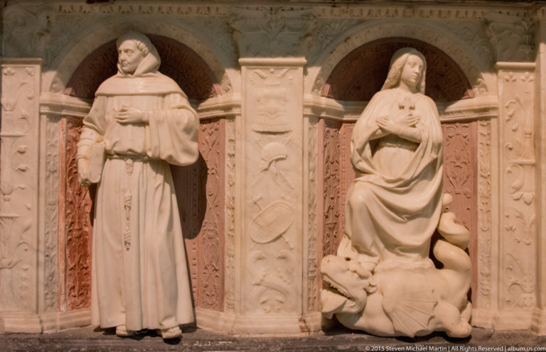 Standing draped female allegorical figure in a carved niche on the side of the ducal tomb, with decorative pilasters and red panels.