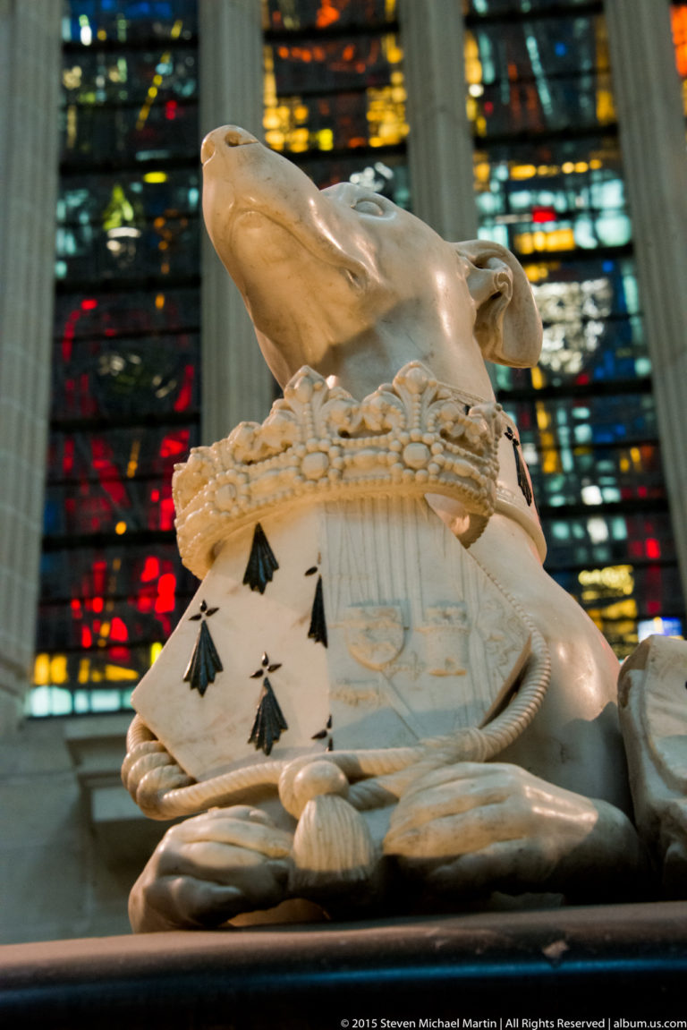 Pale carved heraldic animal bends over a shield patterned with black ermine tails, with stained glass in the background.