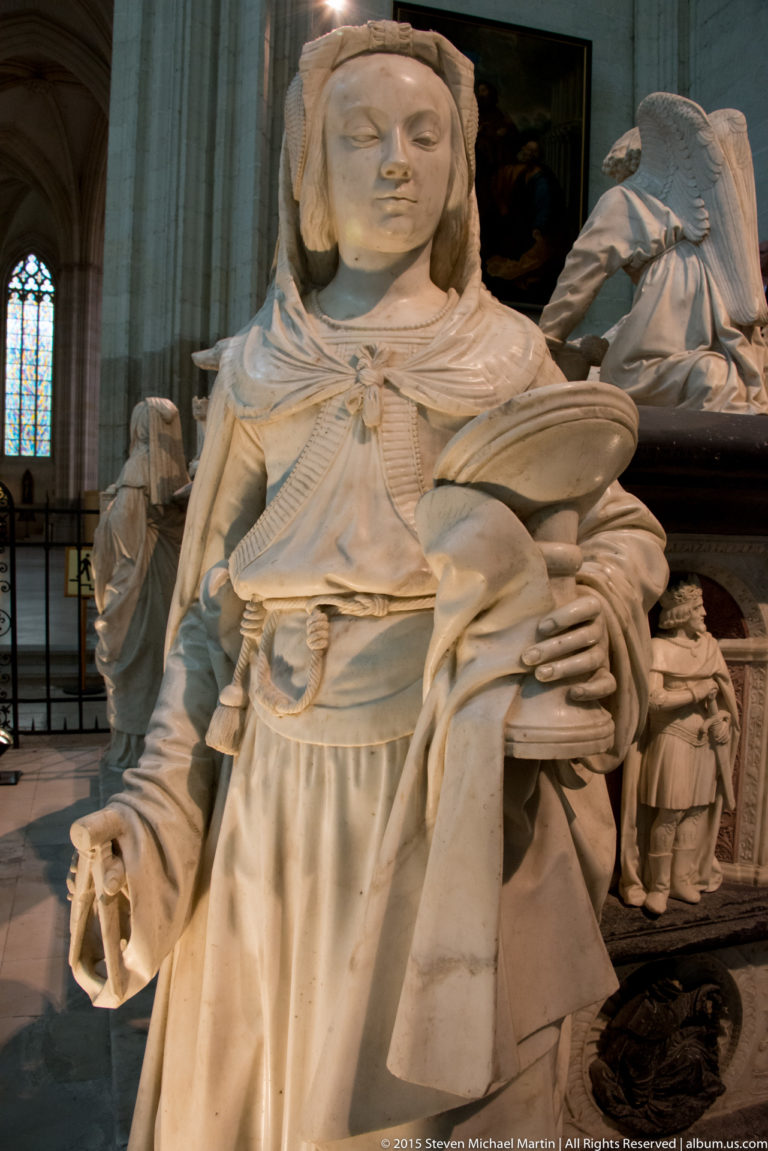 Carved female virtue figure holding an open book, with layered Renaissance dress and neighboring tomb sculptures behind.