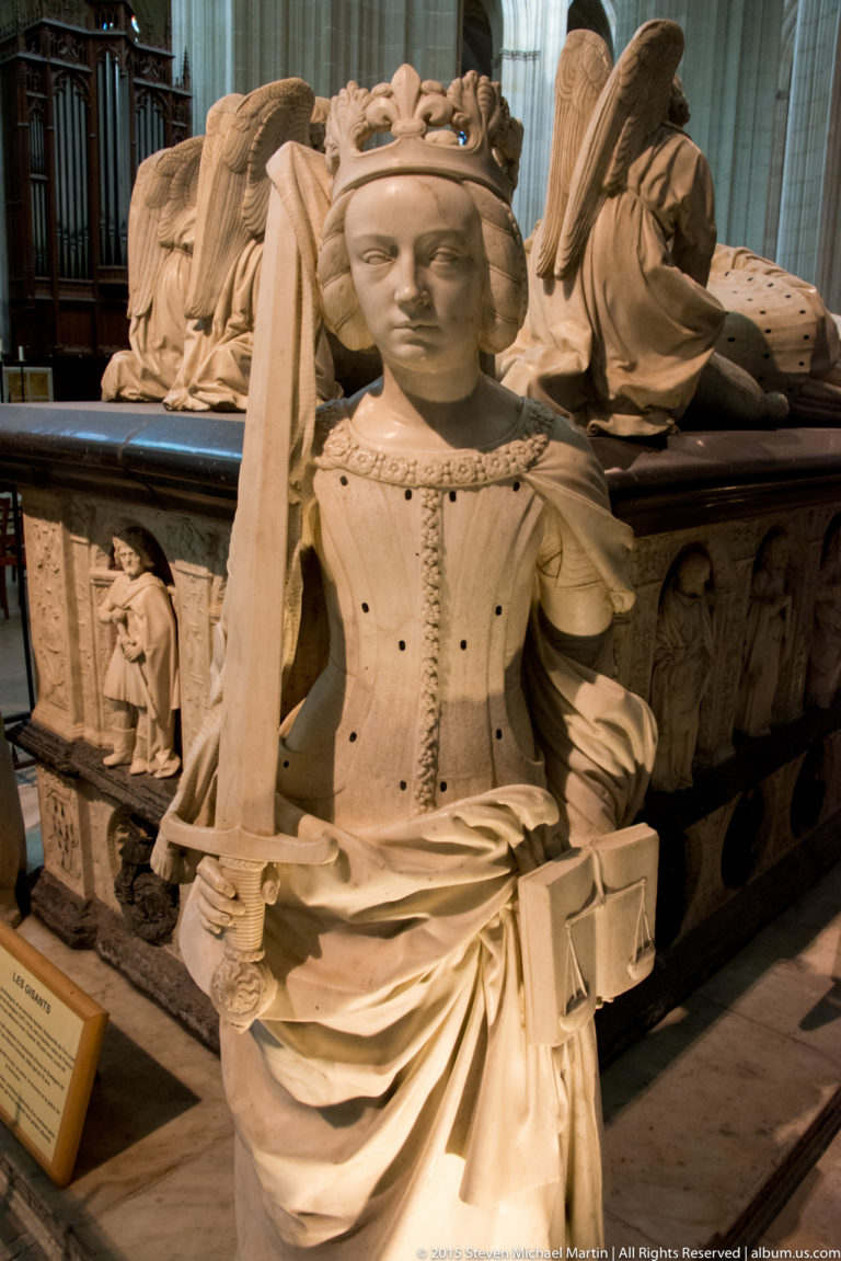 Crowned female virtue figure in carved Renaissance dress holds a long vertical attribute beside the ducal tomb.