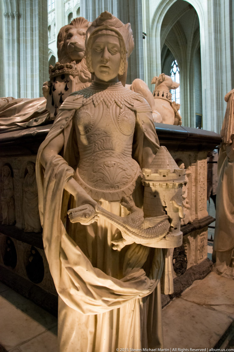 Standing female virtue figure in elaborate carved dress holds a small attribute before the relief-covered side of the ducal tomb.