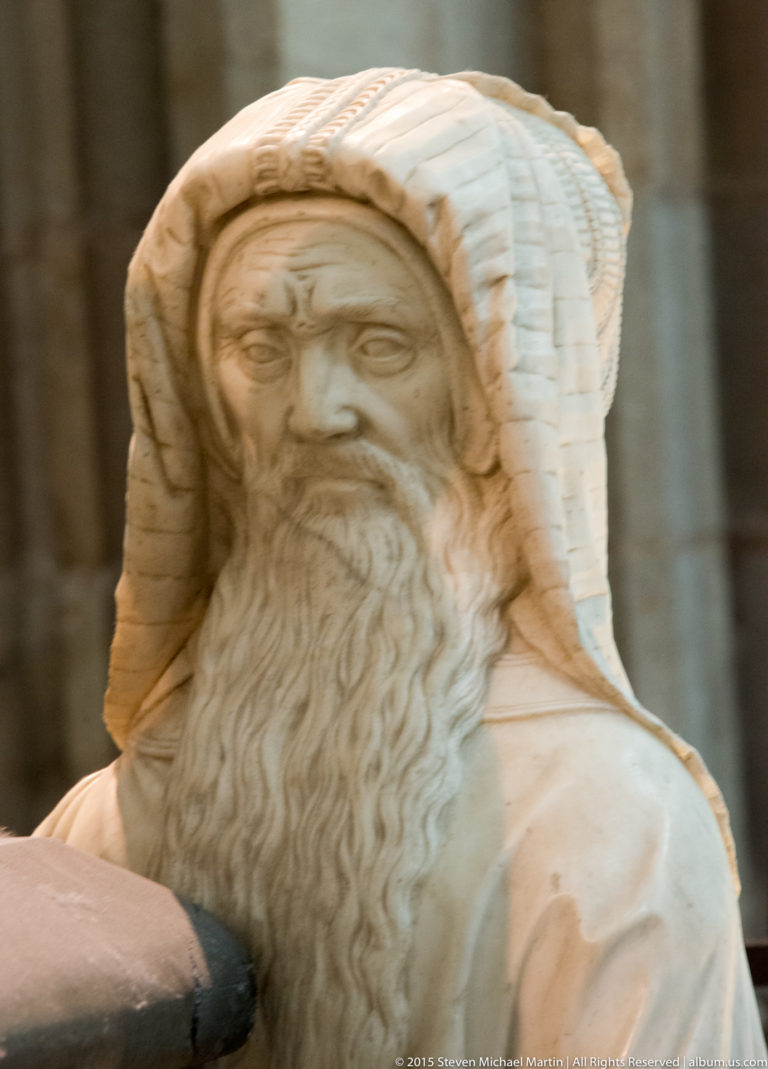 Close view of a carved bearded male head with curled hair, moustache, and folded head covering in pale stone.