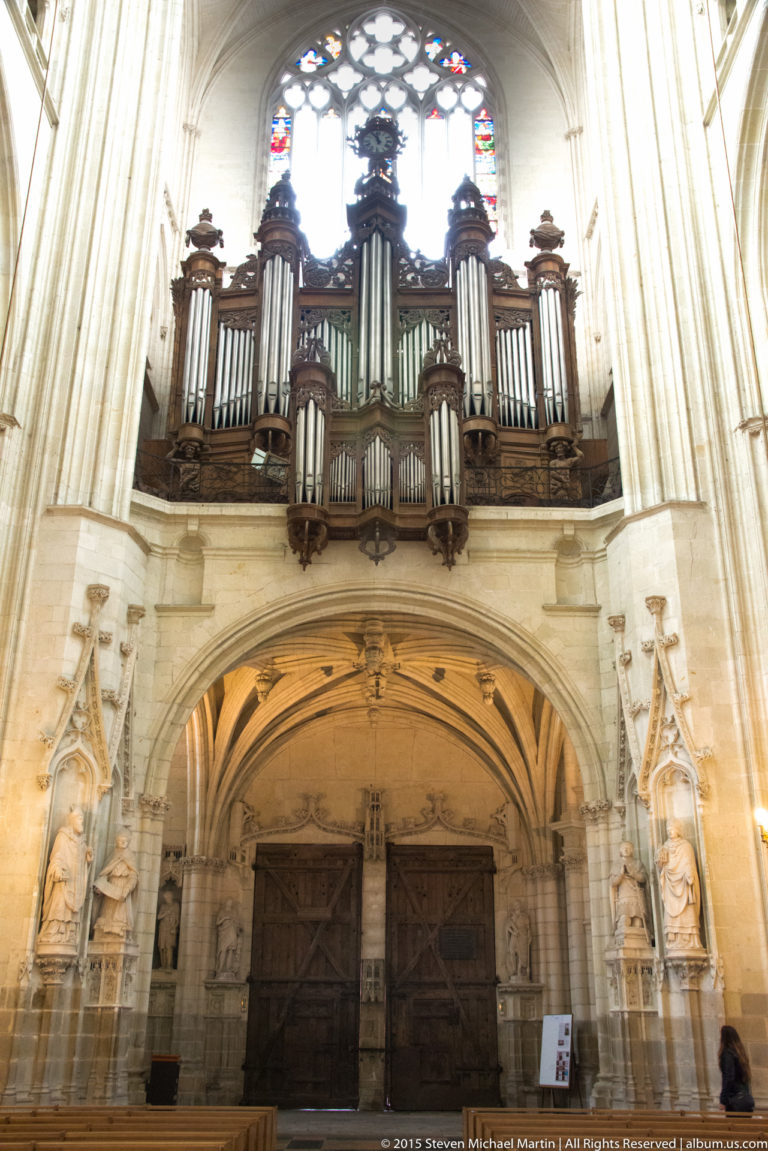 Great organ in a dark carved case above the west doors of Nantes Cathedral, with stained glass and sculpted figures.