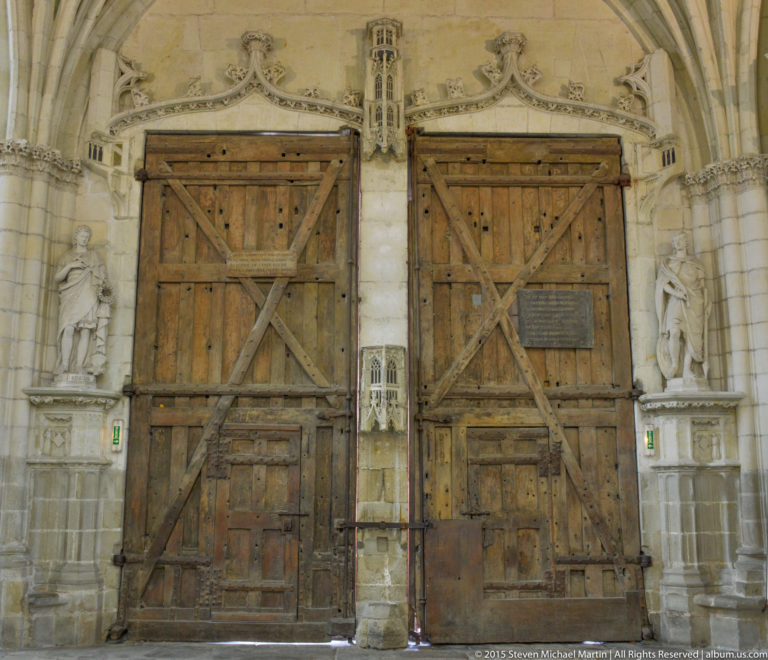 Two massive reinforced wooden doors with diagonal braces and iron fittings beneath carved stone ornament in Nantes Cathedral.