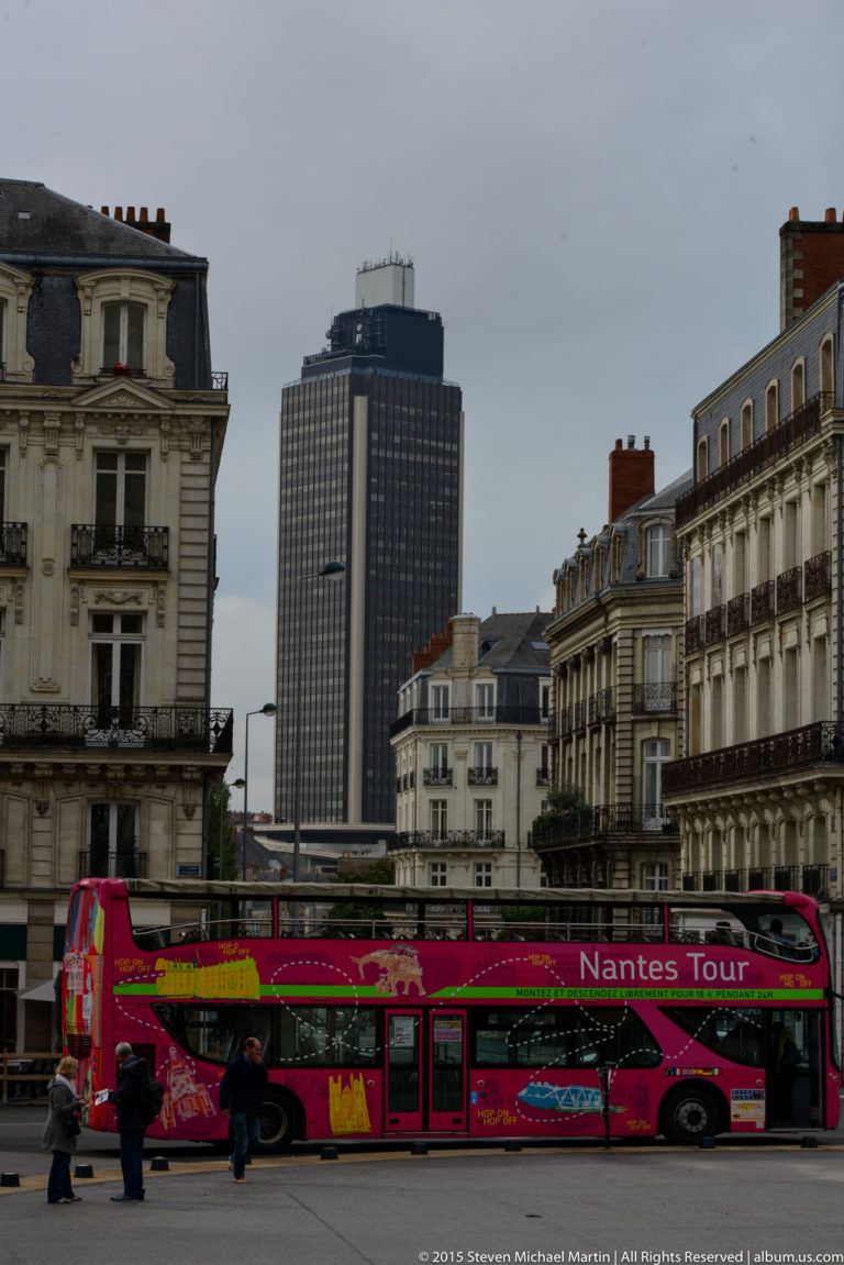 Red Nantes sightseeing bus on a city street with historic façades and the dark high-rise Tour Bretagne behind.