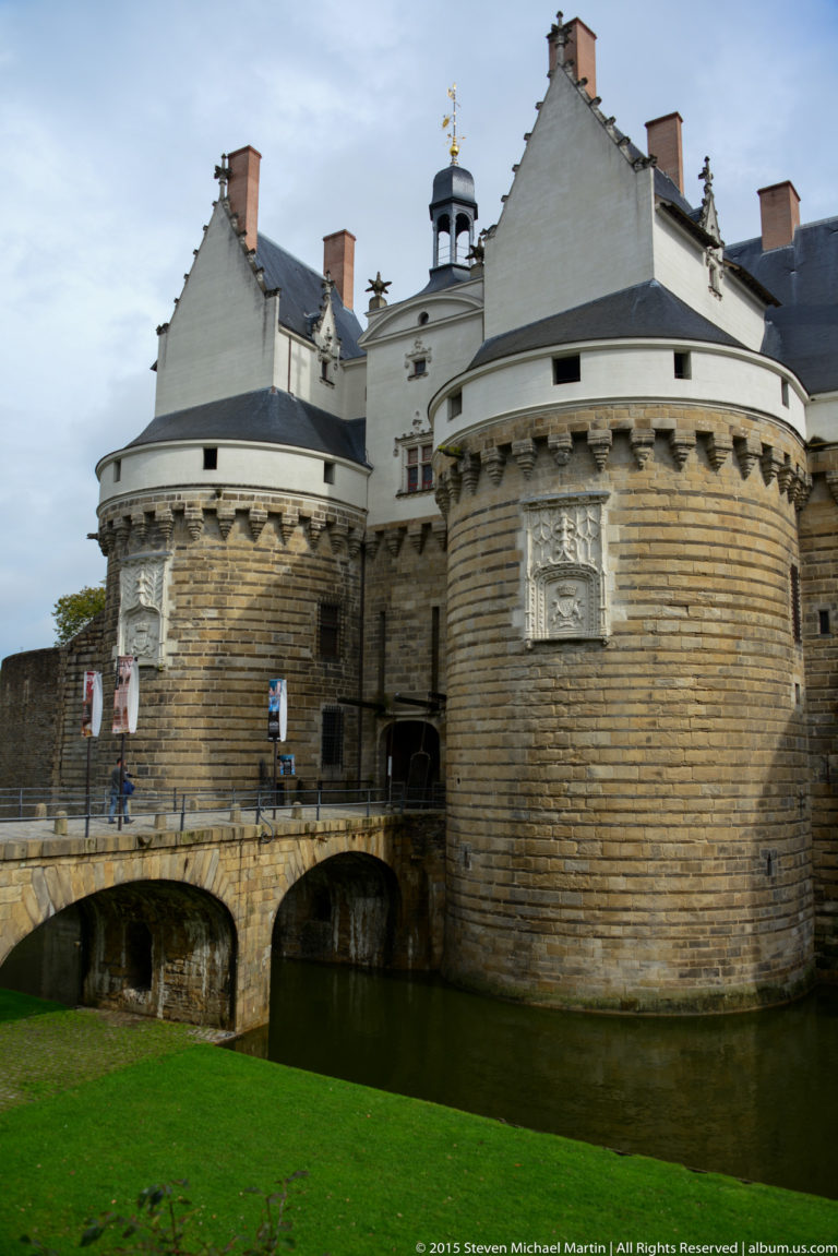 Large round château tower beside the moat, with a low stone bridge and pale residential buildings rising above.