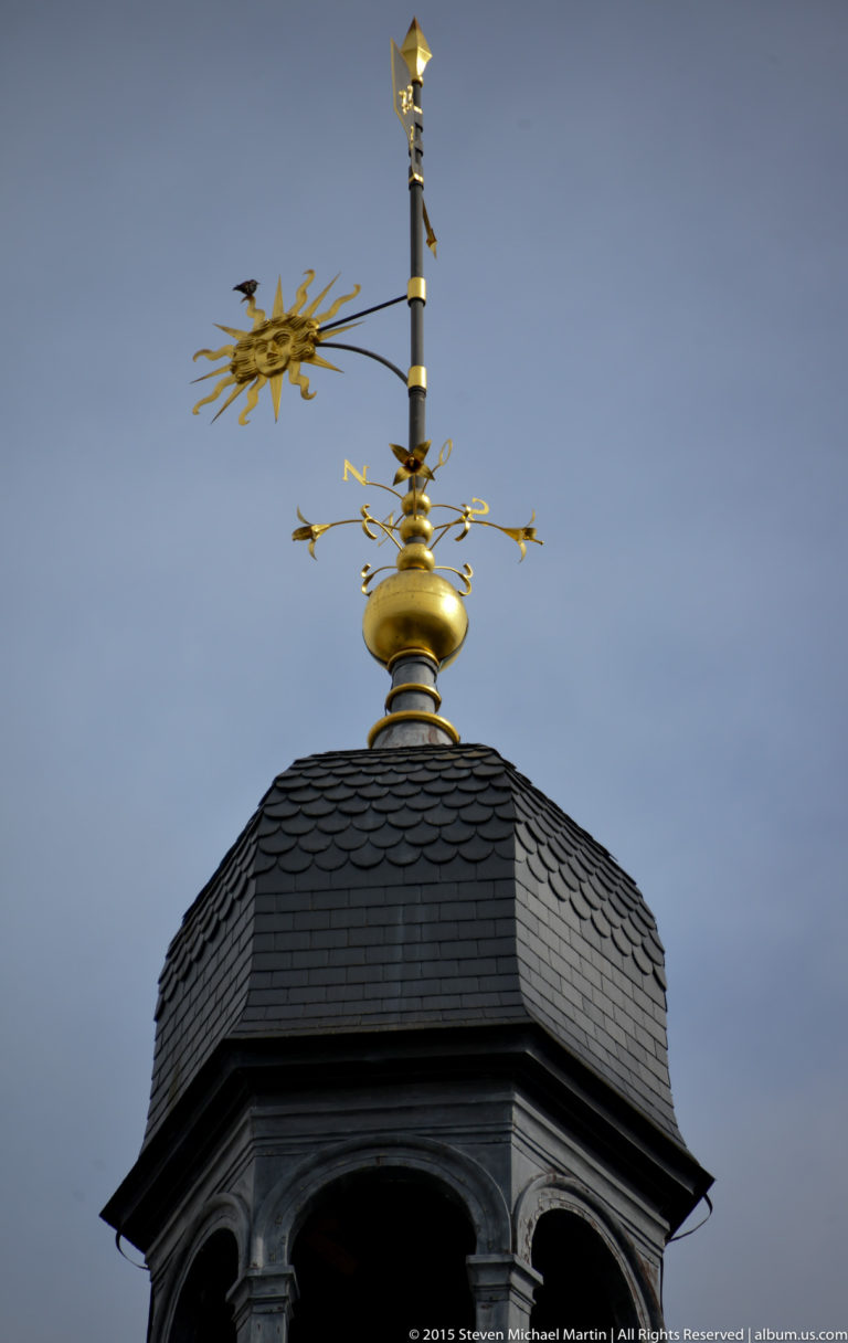 Gilded weather vane and finial above a dark slate turret roof against a gray sky.