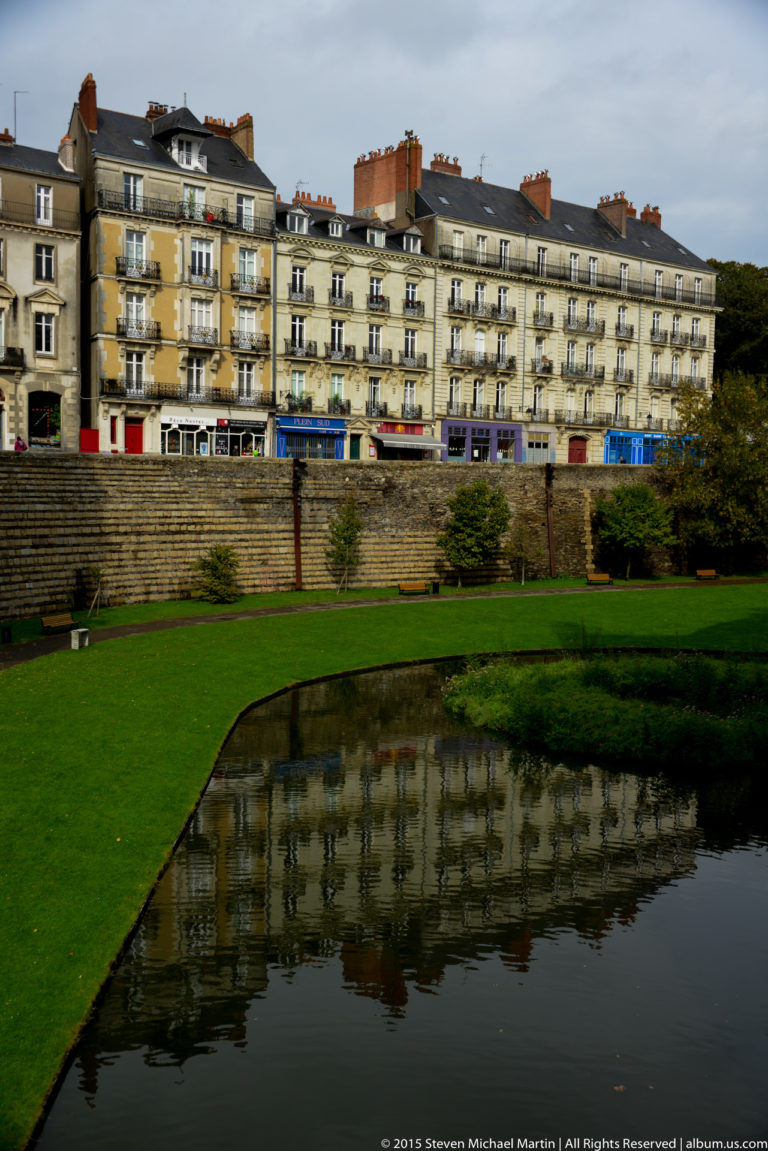 Curving château moat bordered by green lawns, with apartment buildings and defensive walls beyond.