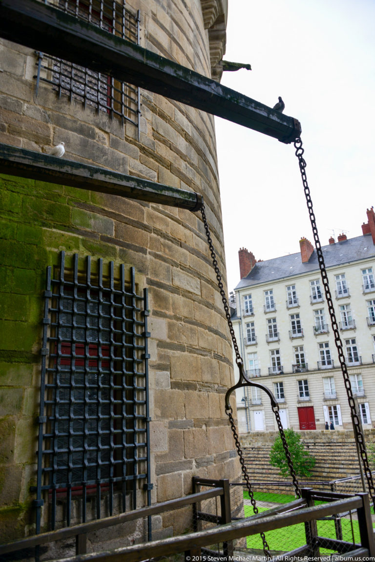 Heavy chains and projecting beams beside a barred opening in the stone wall of the Château des ducs de Bretagne.