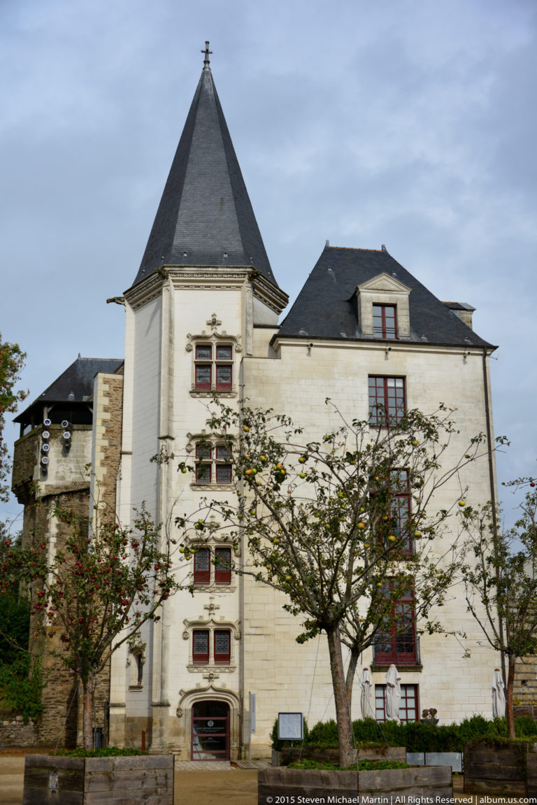 Pale stone château wing with red-framed windows, slate roof, chimney, and square corner tower seen from the courtyard.