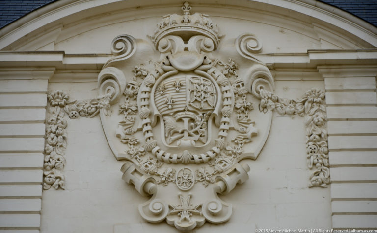 Large carved coat of arms with crown, scrolls, garlands, and pendant insignia on a pale château wall.