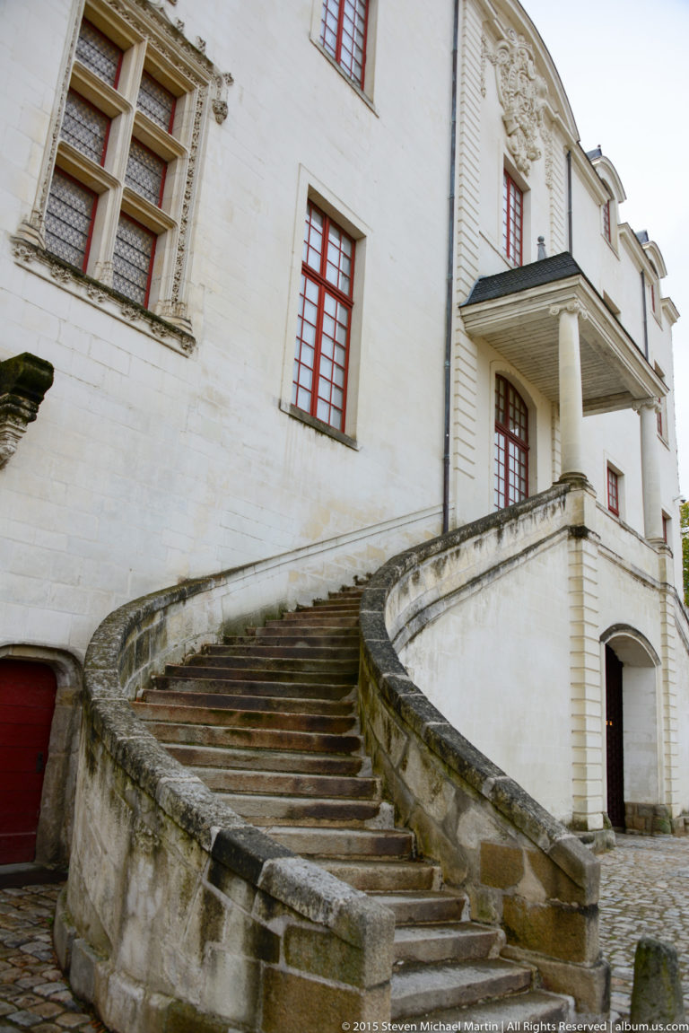 Curving stone staircase rises along a pale château façade with red-framed windows and a small projecting canopy.