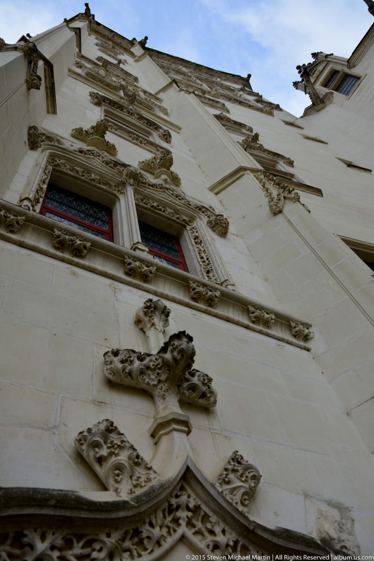 Upward view of a pale château façade with red windows, Gothic tracery, carved foliage, and sculptural stone ornament.