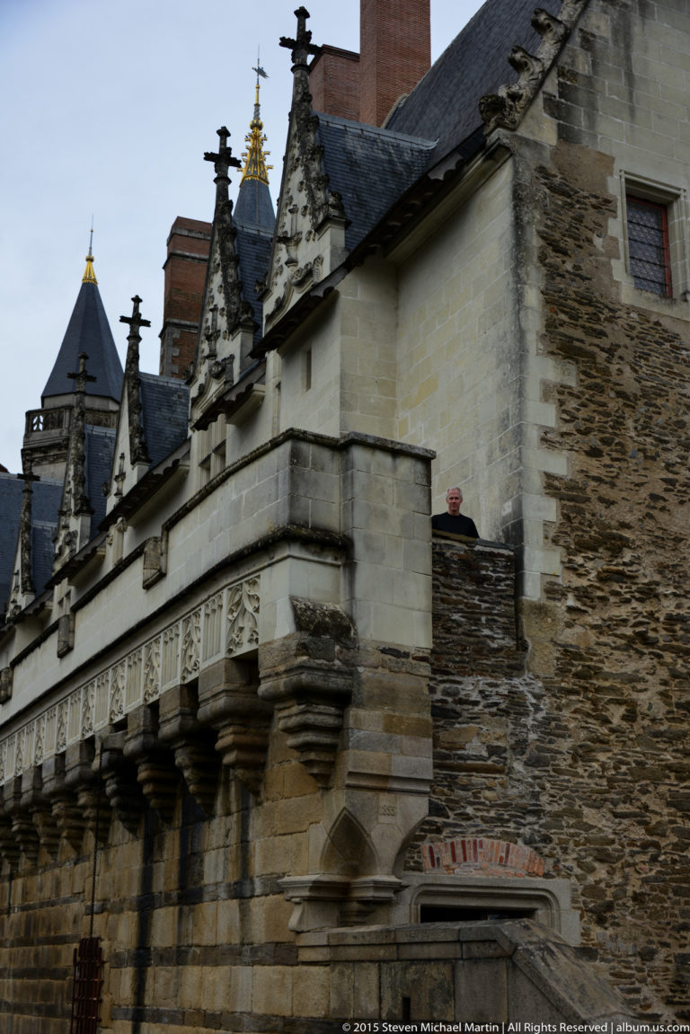 Projecting stone gallery on heavy corbels beside rough château masonry, with stepped gables and chimneys above.