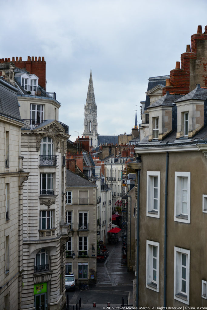 Elevated view down a narrow Nantes street lined with historic buildings, with a tall church tower in the distance.
