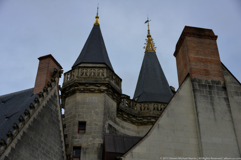 Two stone château turrets with steep slate roofs and gilded finials rise beside tall brick chimneys.