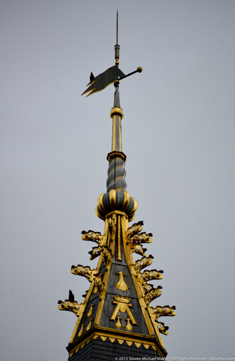 Gilded finial, directional vane, and branching decorative metalwork crown a dark slate château roof.