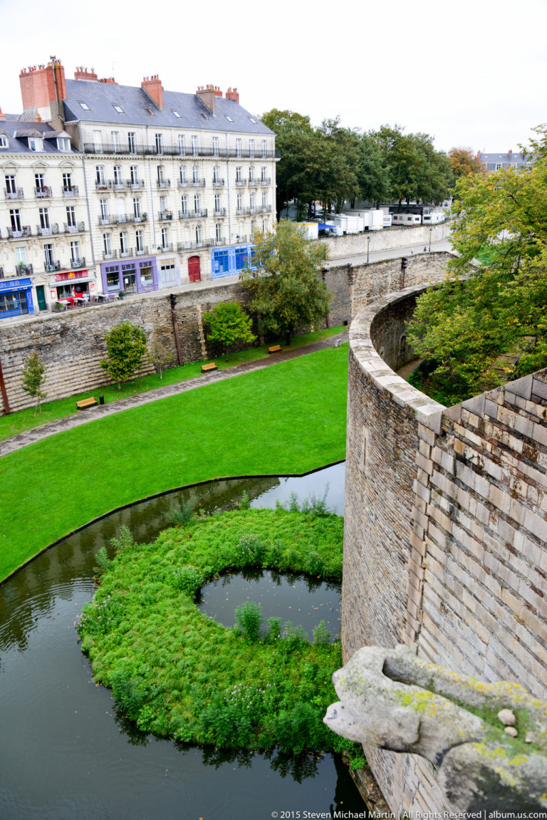Curving stone château wall above a moat with green aquatic plants, lawn, and Nantes buildings beyond.