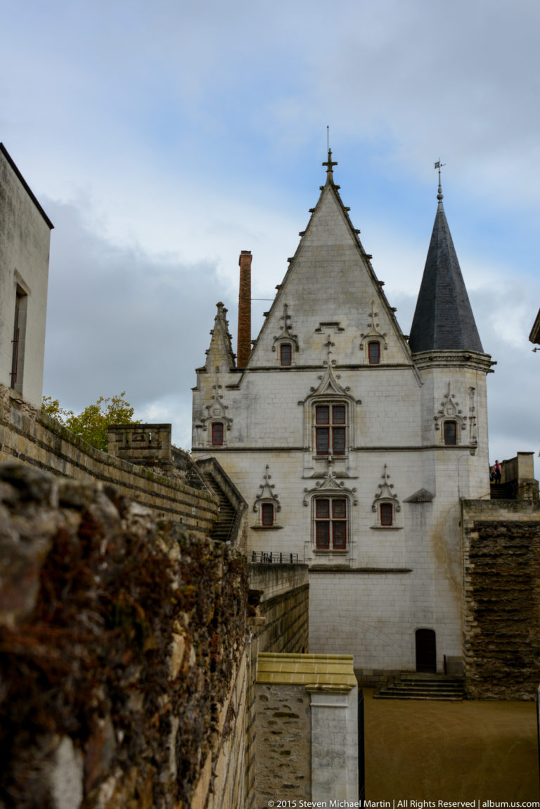 Pale gabled château building with red windows, steep slate roof, chimney, and corner turret seen beyond stone ramparts.