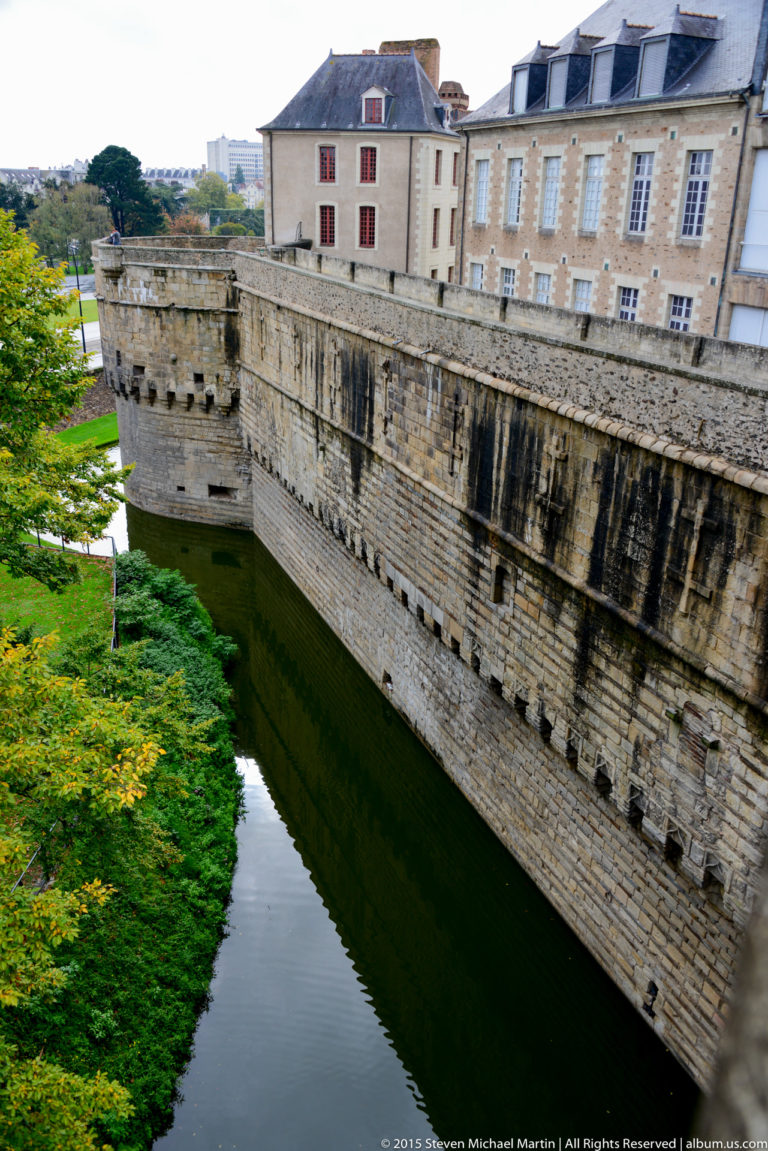 Long weathered stone curtain wall rises from the moat, with château buildings visible above and beyond.