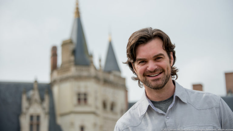 Smiling man in a light gray shirt posed before softly blurred stone château towers with dark slate roofs.
