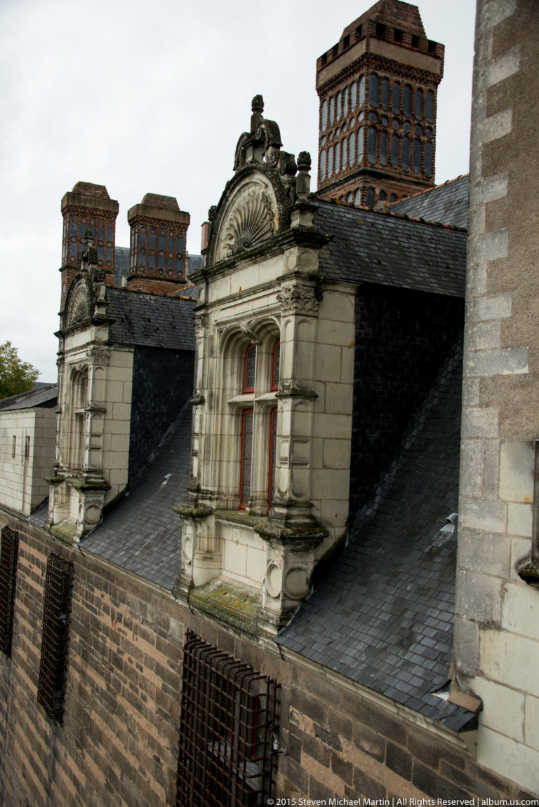 Two ornate stone dormer windows with red frames and tall patterned brick chimneys rising from a dark slate château roof.