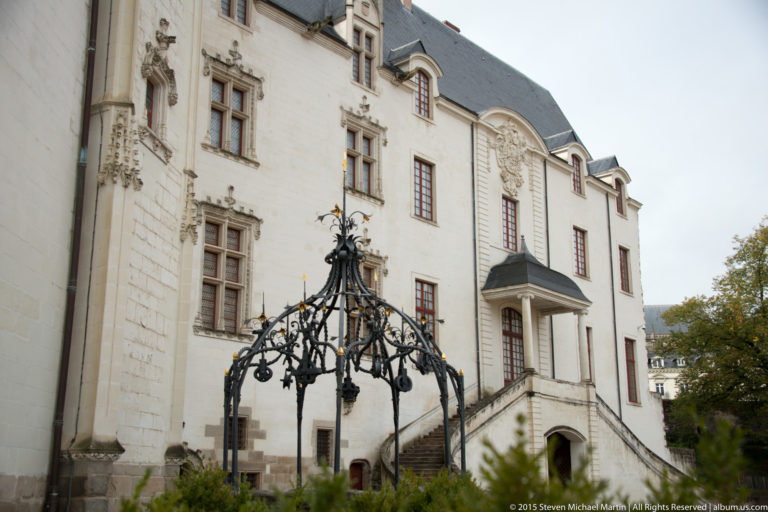 Pale château façade with red-framed windows and exterior stair behind an ornate black wrought-iron structure.