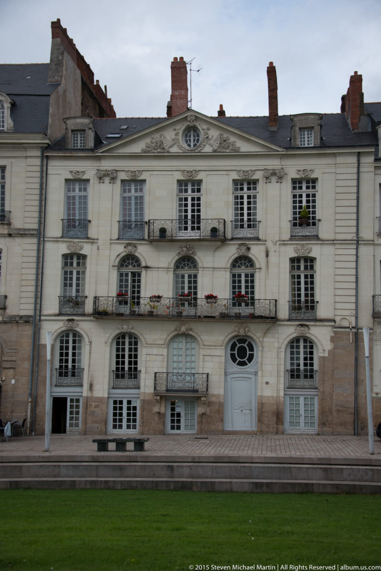 Symmetrical pale historic building with central pediment, oculus, arched ground-floor windows, balconies, and lawn in front.