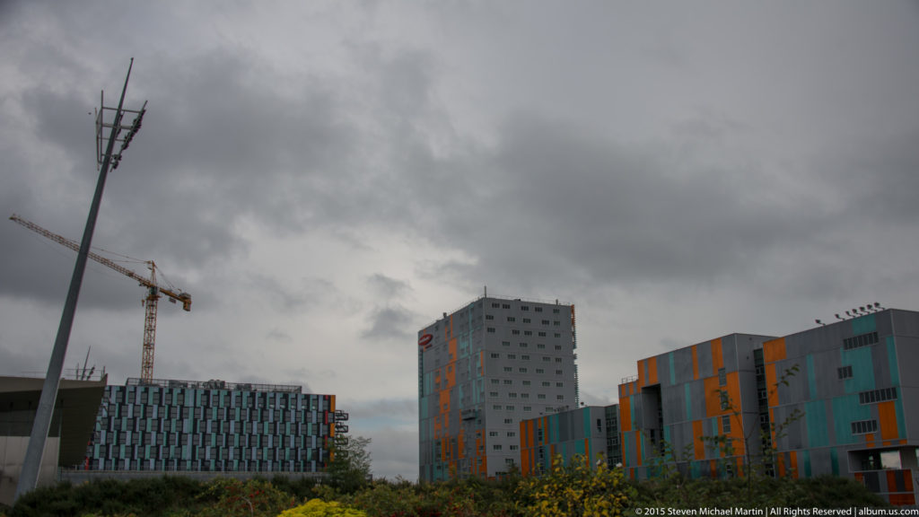 Modern multicolored buildings with teal and orange façade panels beneath dark clouds, with a crane and streetlight nearby.