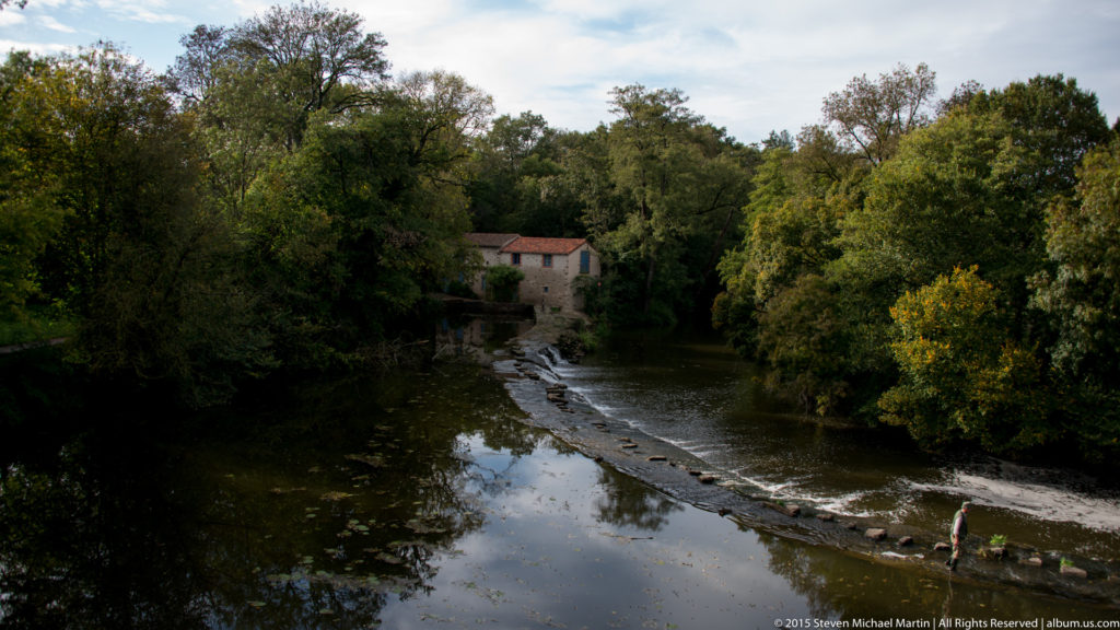 Wooded river with a low weir, reflected trees, and a small building partly hidden on the far bank.