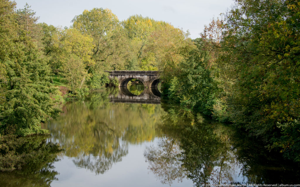 Historic stone bridge with round arches reflected in a calm river surrounded by dense green trees.