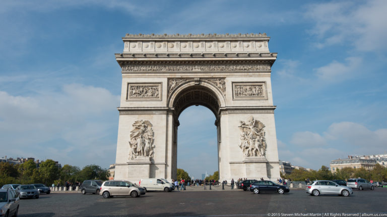 Frontal exterior view of the Arc de Triomphe with its central arch, sculptural groups, blue sky, traffic, and pedestrians.