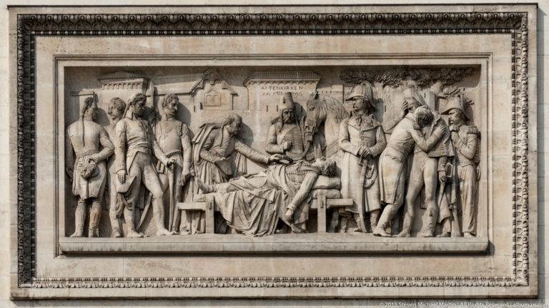 Framed stone bas-relief showing a crowded historical scene with standing figures gathered around a seated central figure.