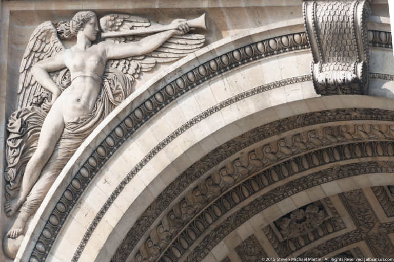 Close upward view of carved arch mouldings, geometric ornament, and part of a monumental stone figure on the Arc de Triomphe.