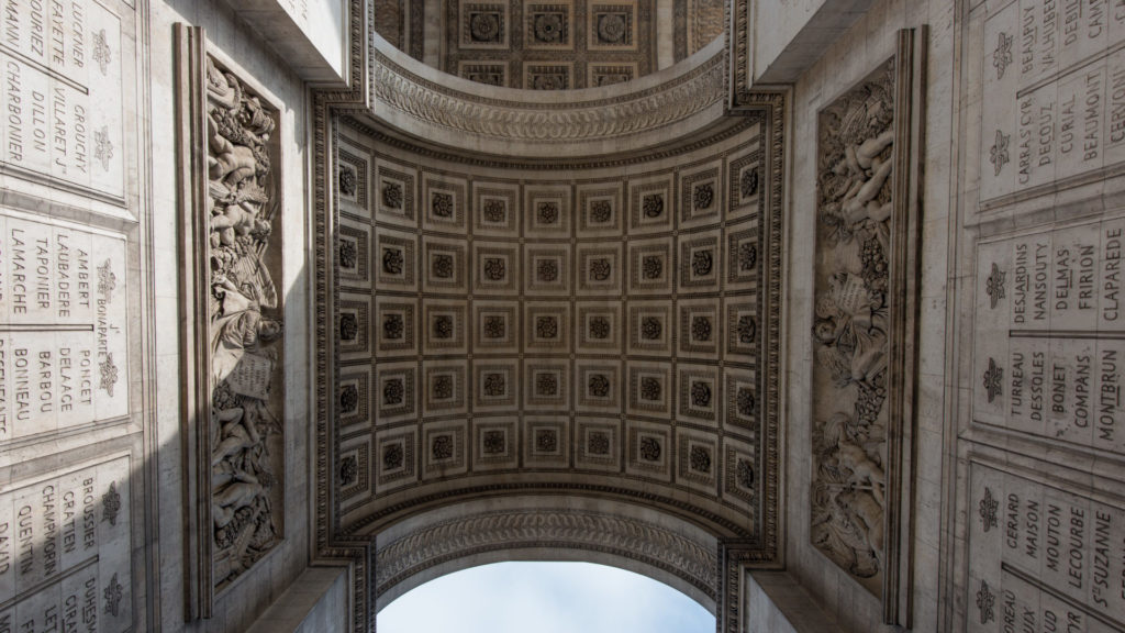 Upward view beneath the Arc de Triomphe showing a coffered barrel vault, inscribed stone piers, and a wedge of sky.