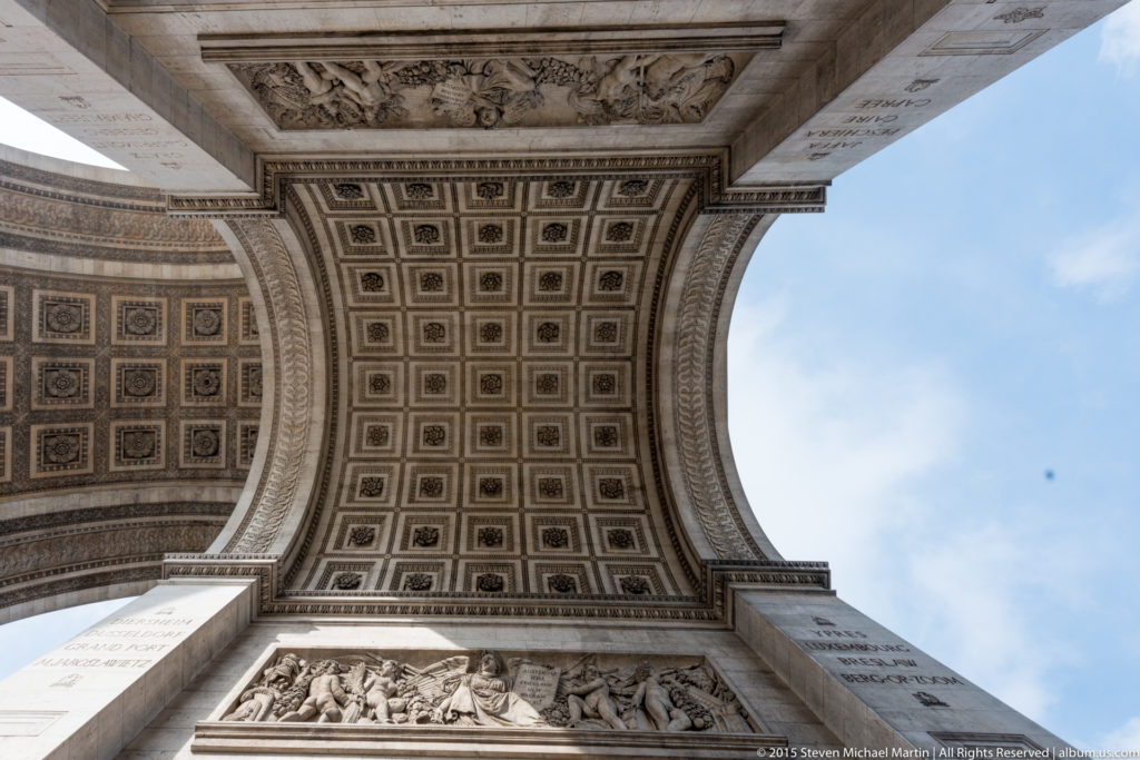 Symmetrical upward view of the Arc de Triomphe’s coffered underside between massive stone piers with blue sky at right.