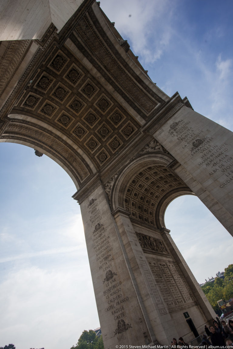 Oblique low-angle view beneath the Arc de Triomphe with diagonal coffered vault, massive stone pier, and open sky.