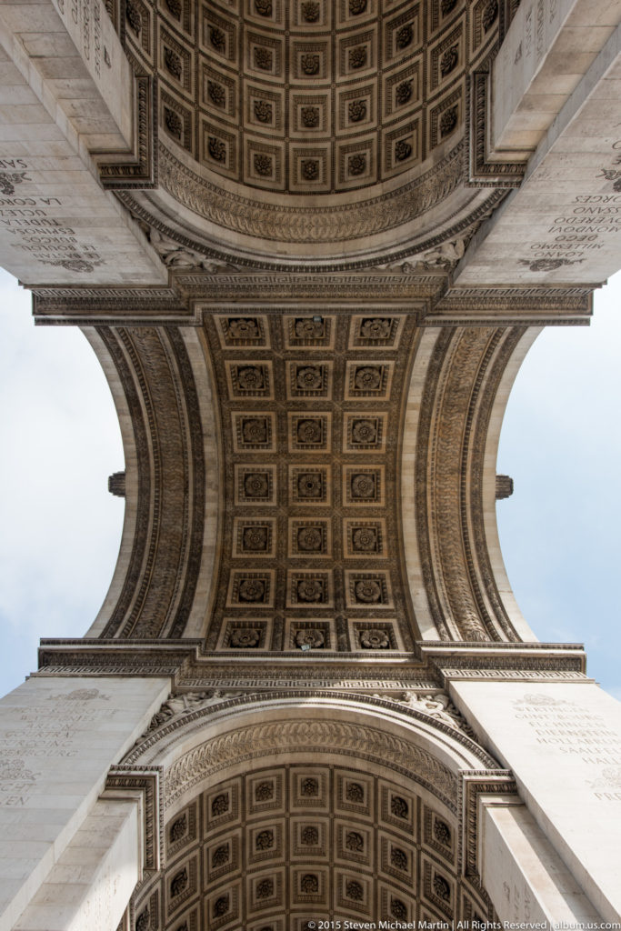 Centered upward view of intersecting coffered arches and stone mouldings beneath the Arc de Triomphe.