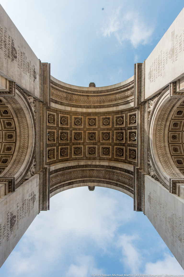 Wide upward view of the Arc de Triomphe’s coffered vault between inscribed stone piers under a blue sky.