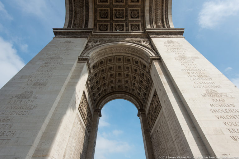 Vertical low-angle view between inscribed stone piers toward the coffered arch and blue sky of the Arc de Triomphe.