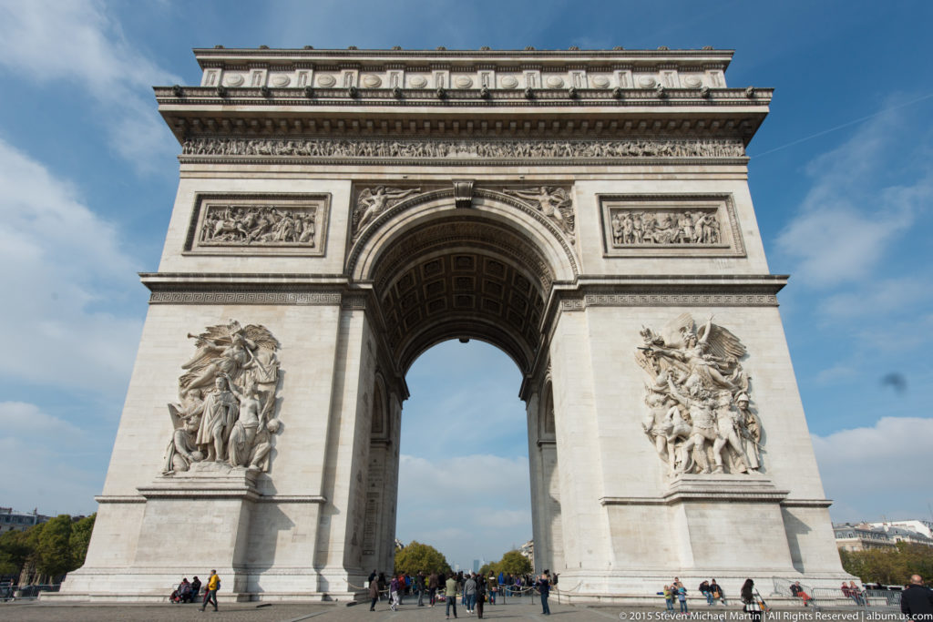 Centered full-height view of the Arc de Triomphe with paired pier sculptures, large central opening, pedestrians, and blue sky.