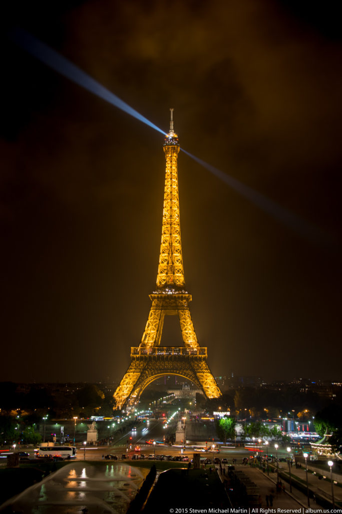 Illuminated Eiffel Tower centered above the Champ de Mars at night, with a searchlight beam crossing the cloudy sky.