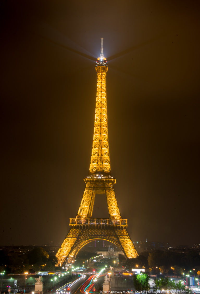 Illuminated Eiffel Tower at night above red and white traffic light trails, with a bright searchlight at its summit.