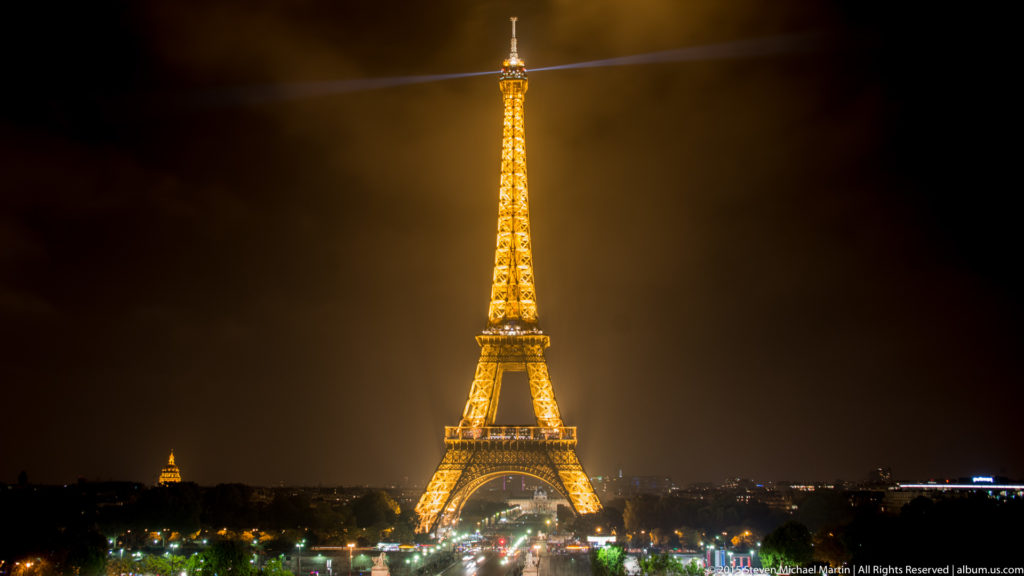 Full-height Eiffel Tower glowing gold at night, vertically framed above a dark Paris skyline with its searchlight extending sideways.