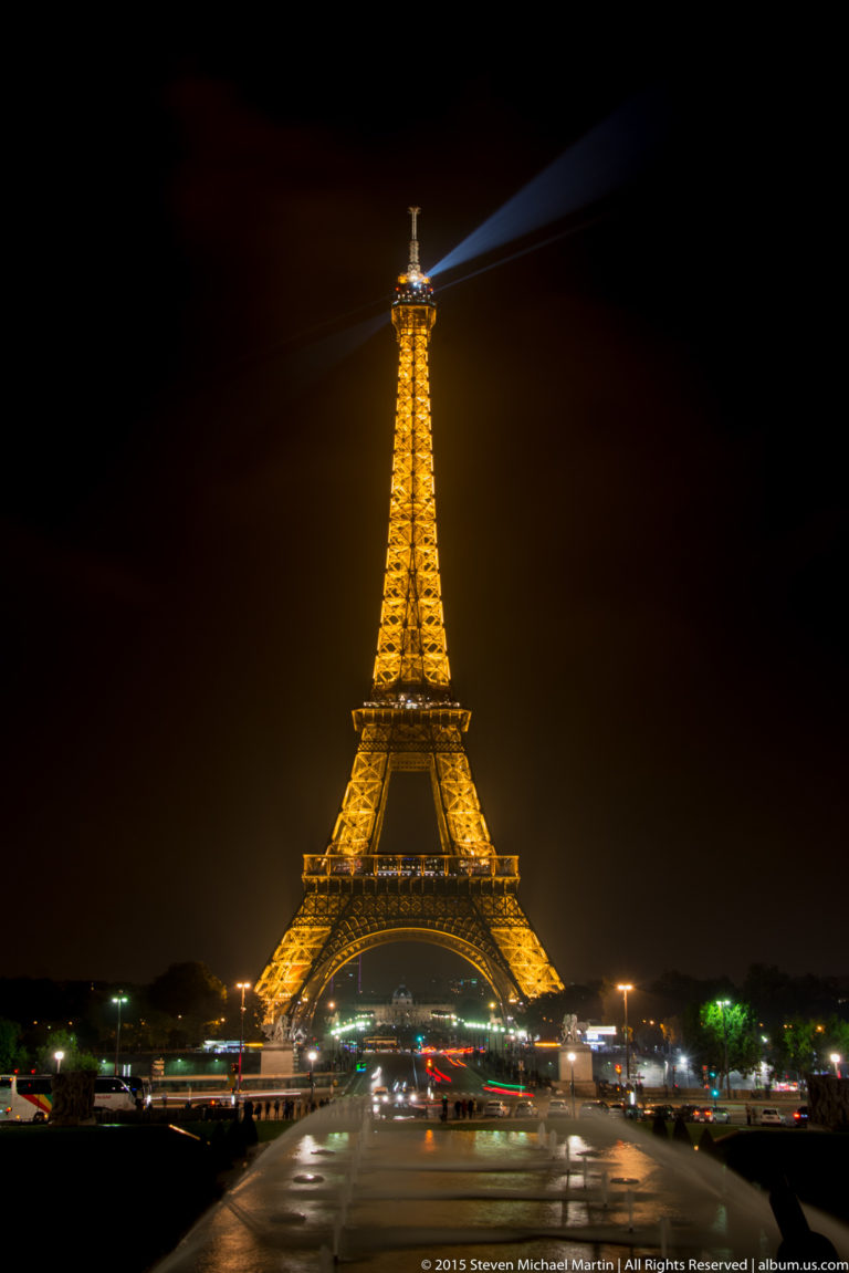 Golden Eiffel Tower viewed symmetrically across the dark Champ de Mars at night, with a searchlight cutting across the sky.