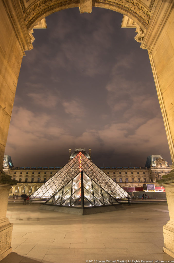Illuminated Louvre Pyramid at night framed beneath a large stone palace arch in the Cour Napoléon.